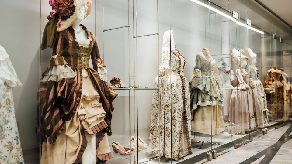 An elegant display of mannequins dressed in intricate, vintage gowns from various historical periods, showcased behind glass in a museum setting.