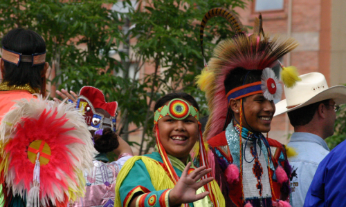 A vibrant scene of a traditional celebration, featuring participants in colourful and elaborate Indigenous attire, smiling and engaging in the festivities.