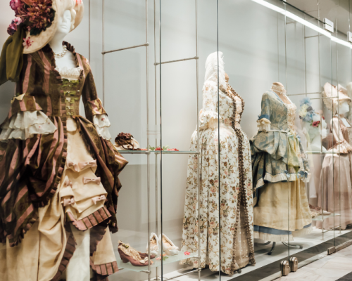 An elegant display of mannequins dressed in intricate, vintage gowns from various historical periods, showcased behind glass in a museum setting.