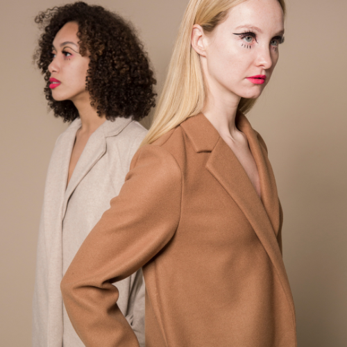 Two women stand back-to-back, showcasing stylish, neutral-toned coats against a plain background.