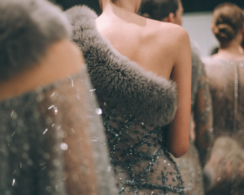 A line of models wearing elegant, fur-trimmed gowns stand backstage, showcasing intricate designs and luxurious fabrics.